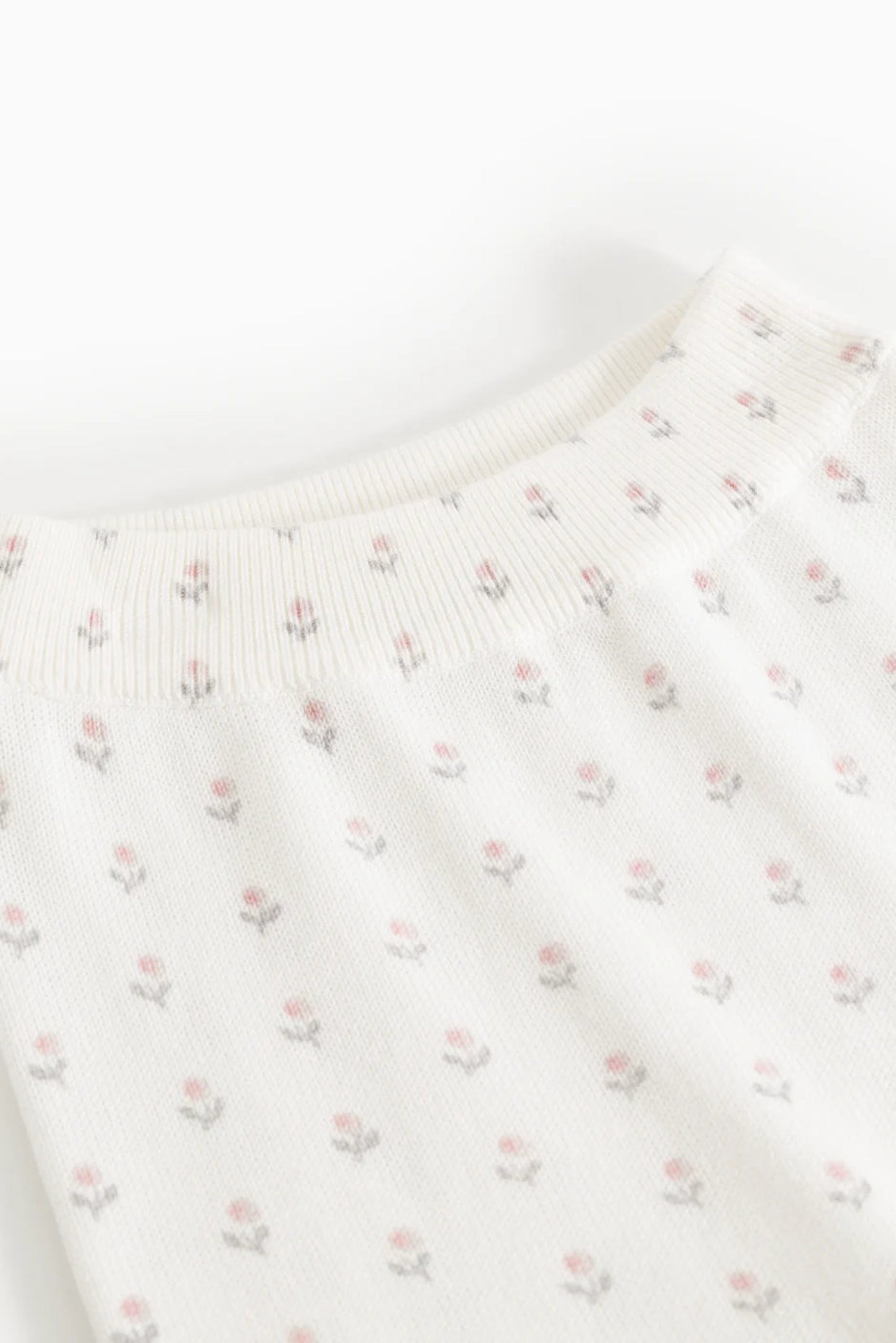 Close-up of a cream-colored knit garment with a delicate all-over small floral print in soft pink and muted gray-green, showing the ribbed crew neck.