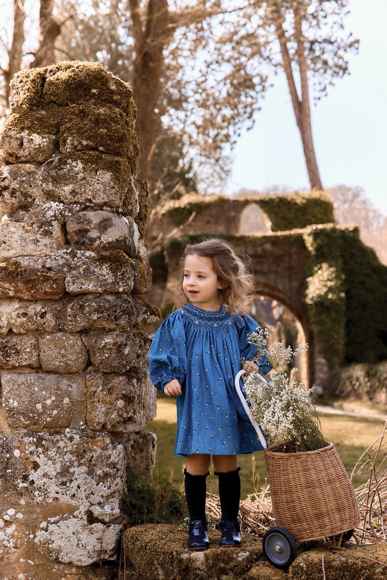 A young girl stands outdoors in front of a mossy stone wall, wearing a blue long-sleeve dress with small white star-like details, black knee-high socks, and dark rain boots while holding a wicker basket filled with dried flowers.