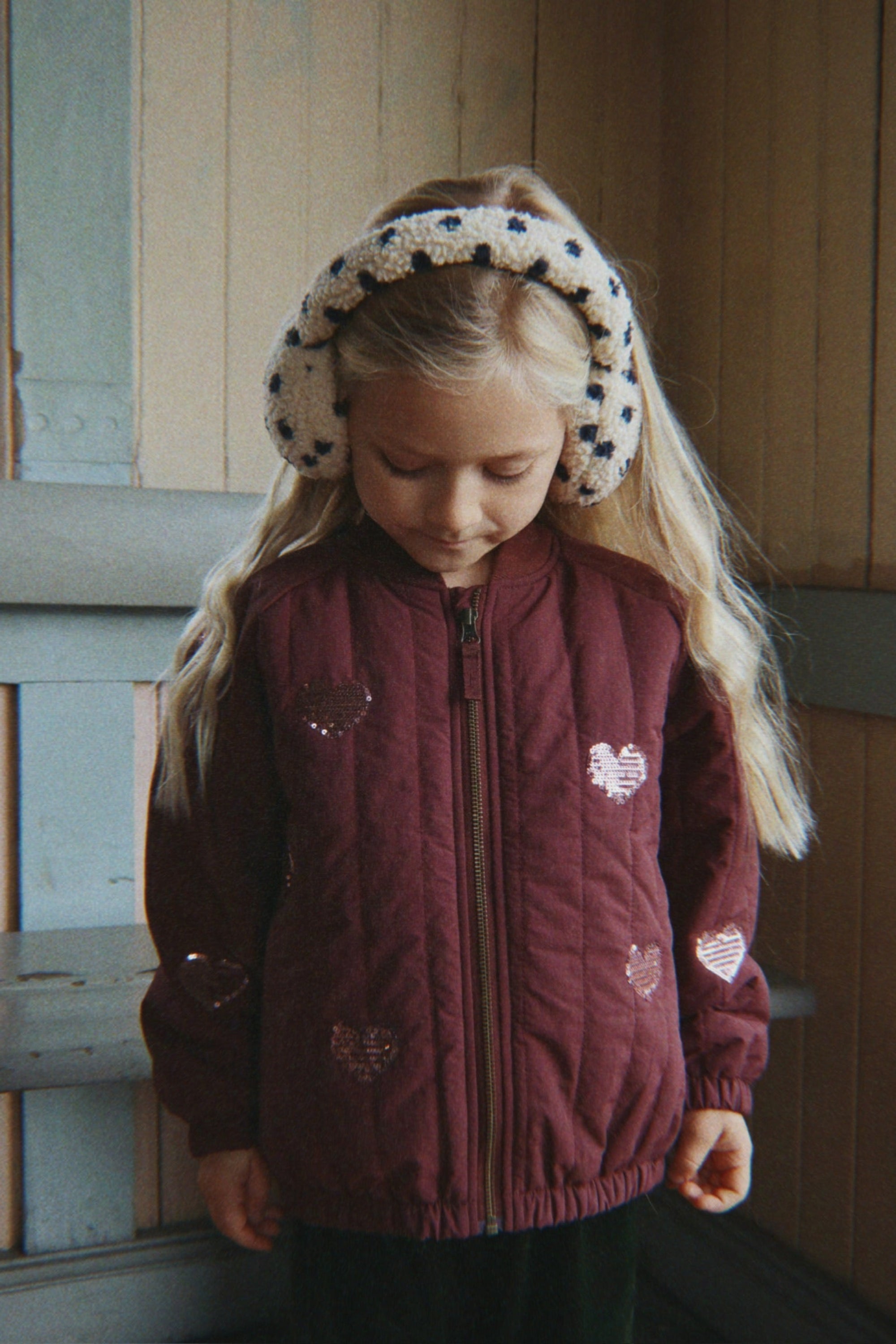 Child wearing a maroon quilted jacket with heart patterns and white earmuffs with black polka dots.