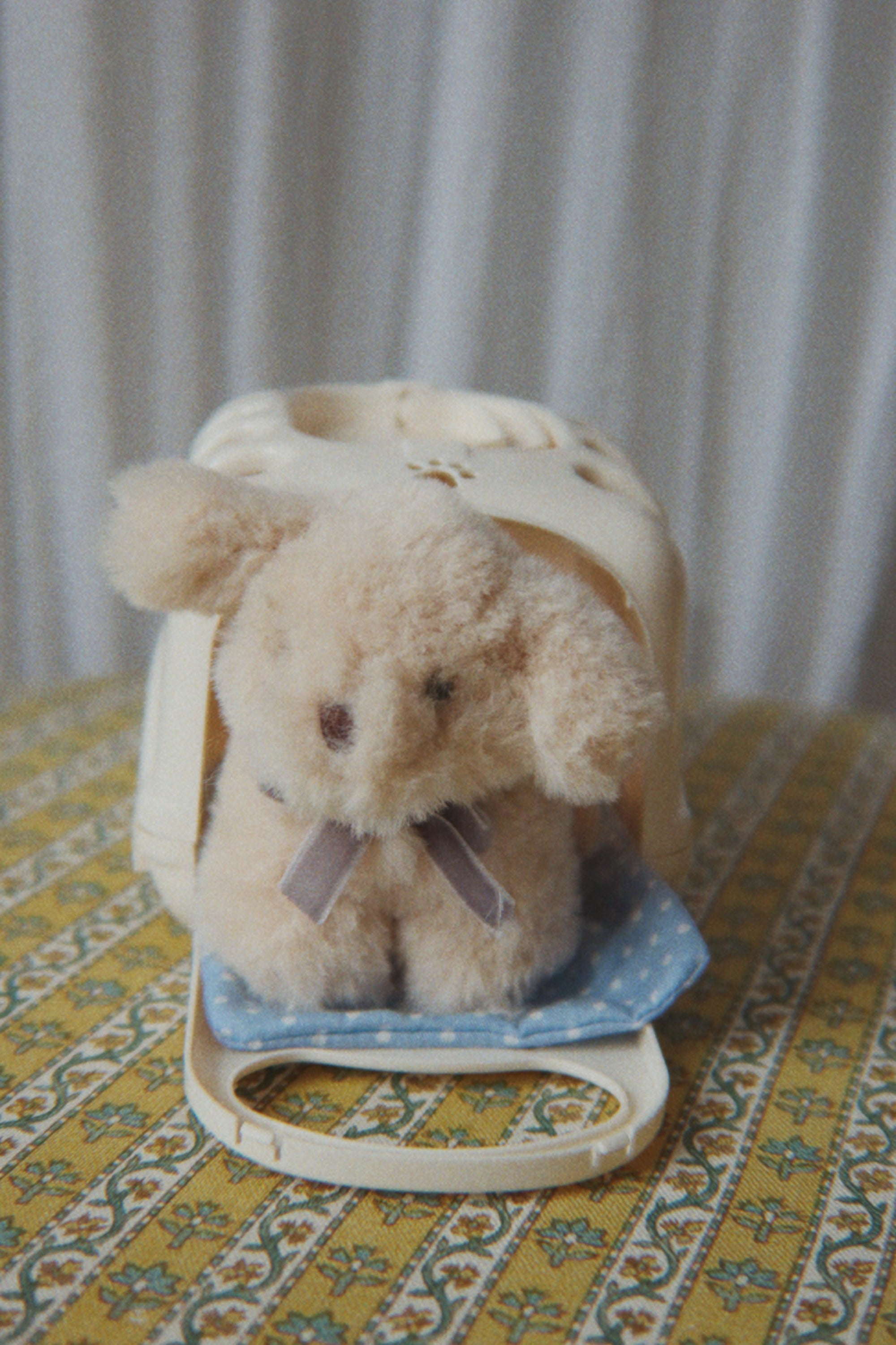 Small plush toy with a bow on a decorative stand against a white curtain background